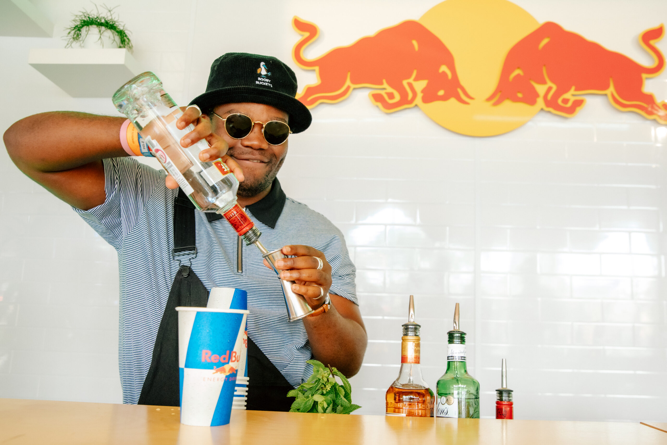 A bartender in a black hat and sunglasses pours from a vodka bottle into a spirit jigger with a Red Bull branded cup in the foreground and the Red Bull logo behind. From the Red Bull Lab Bar activation at Latitude Festival 2021