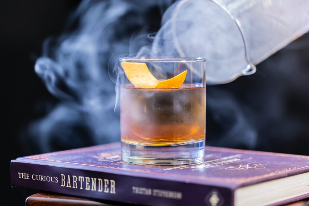 An old fashioned cocktail in a rocks glass is shrouded in smoke