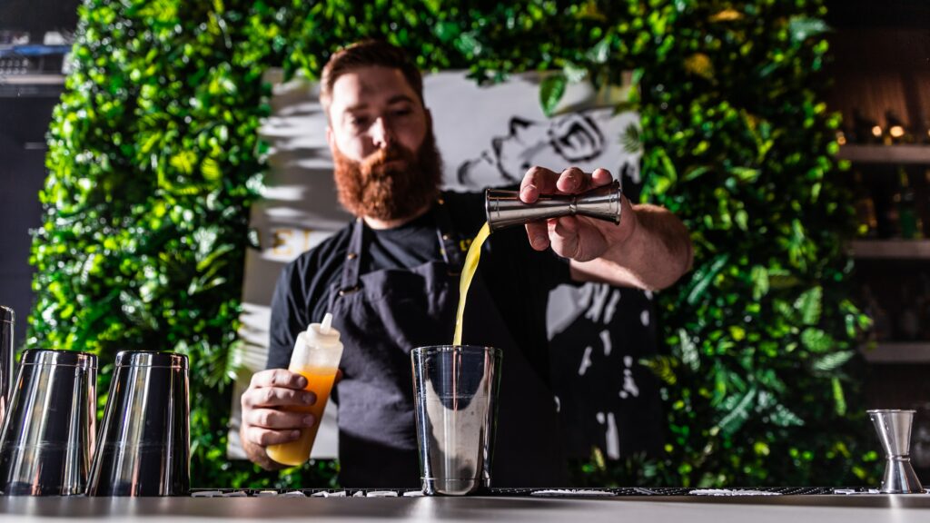A mixologist at an event pours fruit juice into a cocktail shaker