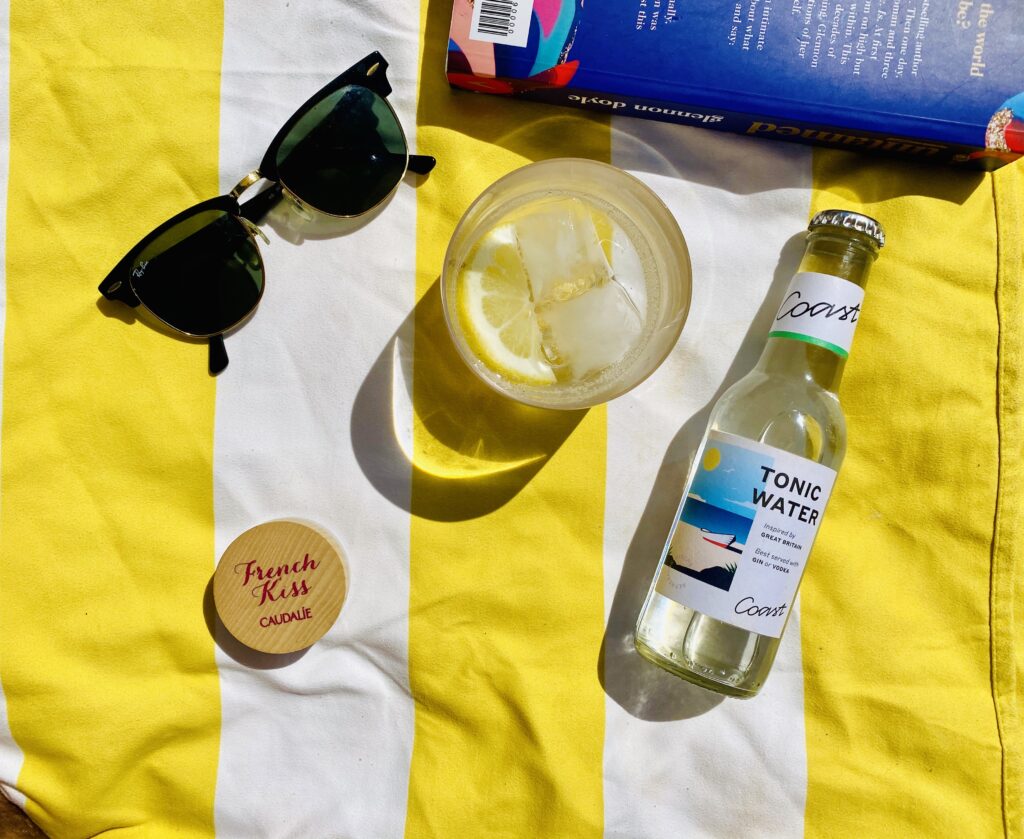 COAST Drinks tonic next to a cocktail and sunglasses, sat on a beach towel