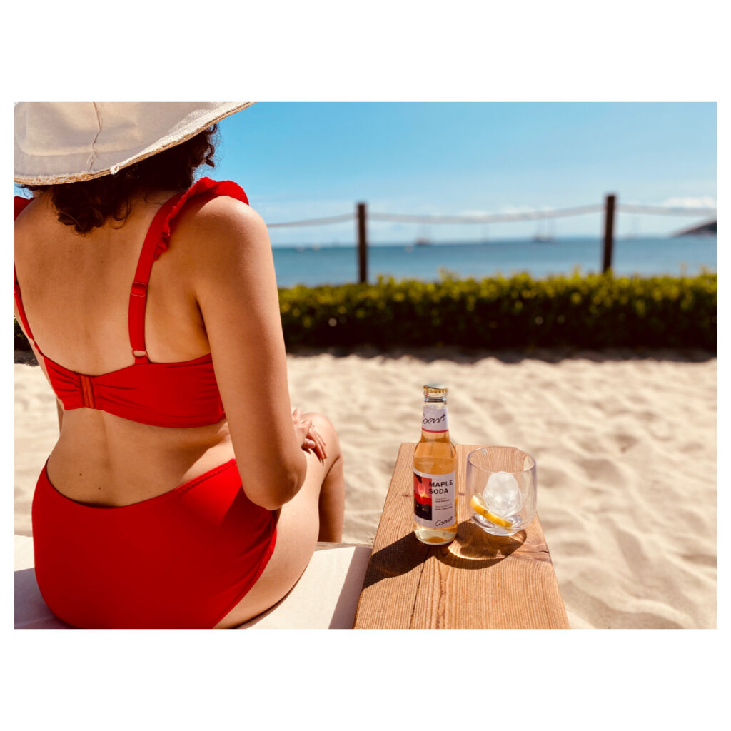 A woman in a red bikin sits on the beach next to a COAST Drinks mixer bottle